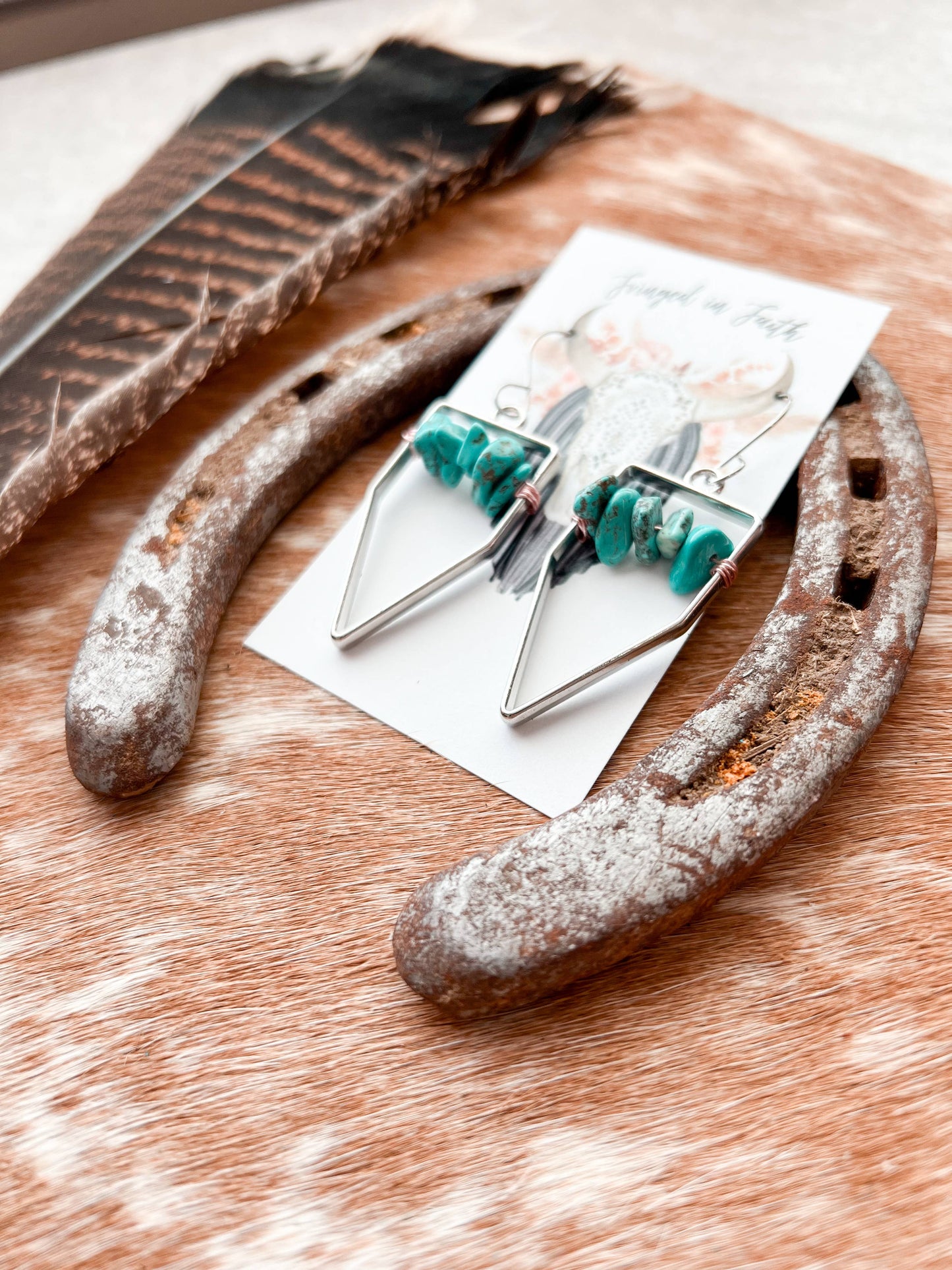 Boho-style earrings with turquoise beads and silver wire elements, displayed on a white card with rustic props including a feather and a horseshoe on a cowhide surface.
