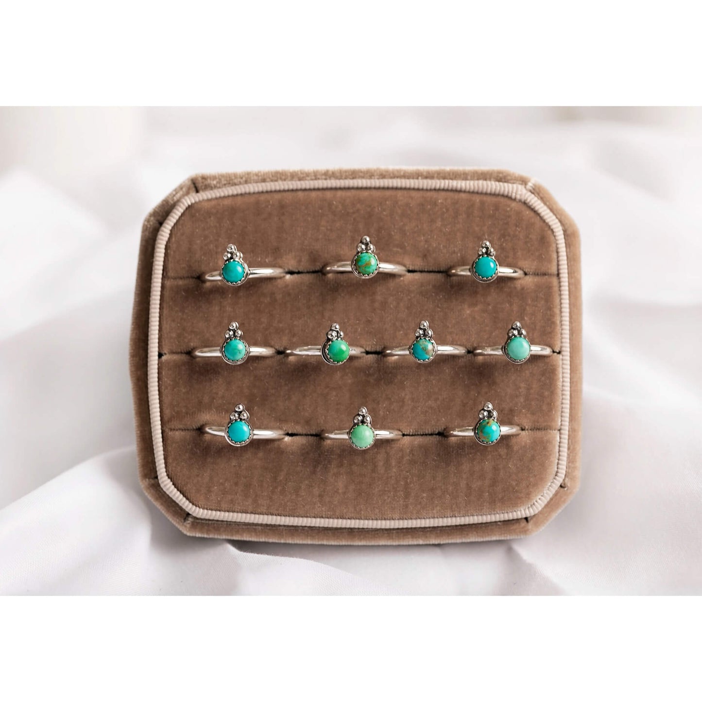 A set of nine silver rings with small oval turquoise-blue stones, displayed on a brown velvet ring box against a white fabric background.