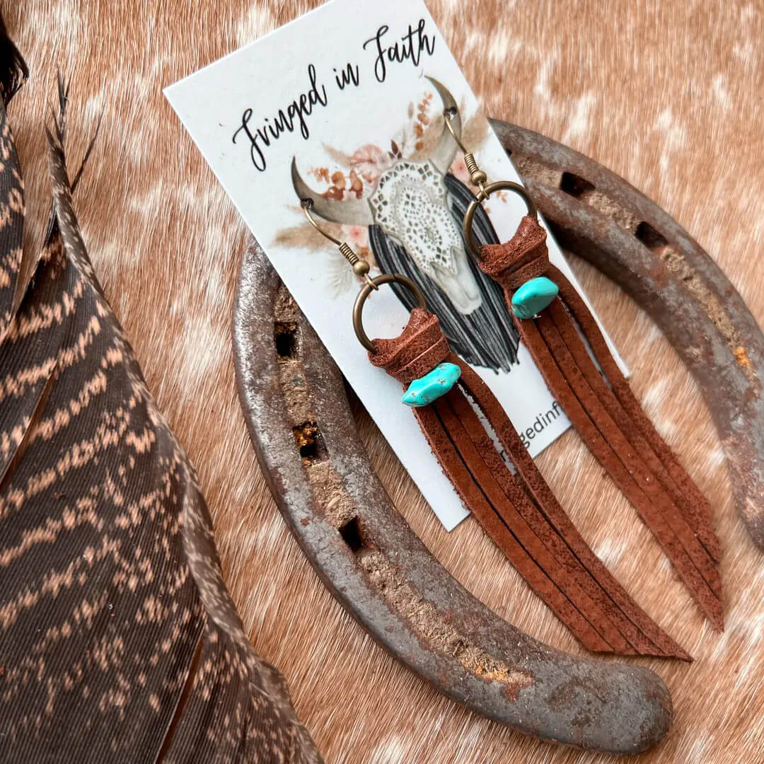 Boho-style dangle earrings displayed on a branded card, styled on a rustic metal horseshoe over a cowhide surface with a feather at the side. The earrings have brown leather fringe, gold-toned rings, a feather accent, and turquoise-colored beads.