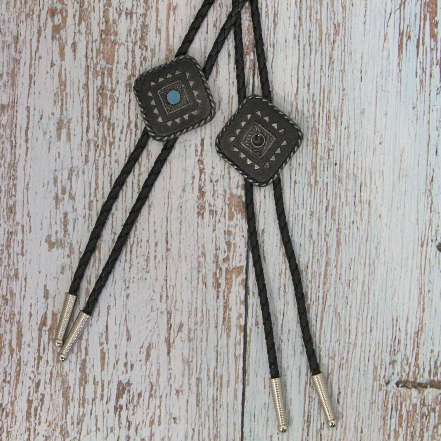 Two diamond-shaped bolo ties with Aztec design, featuring German silver and black stone, on a black braid against a wooden background.