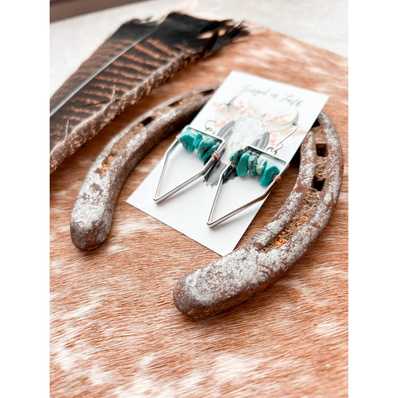Silver geometric dangle earrings with turquoise beads, displayed on a white card and placed on a rustic brown surface with a feather and metal piece nearby.