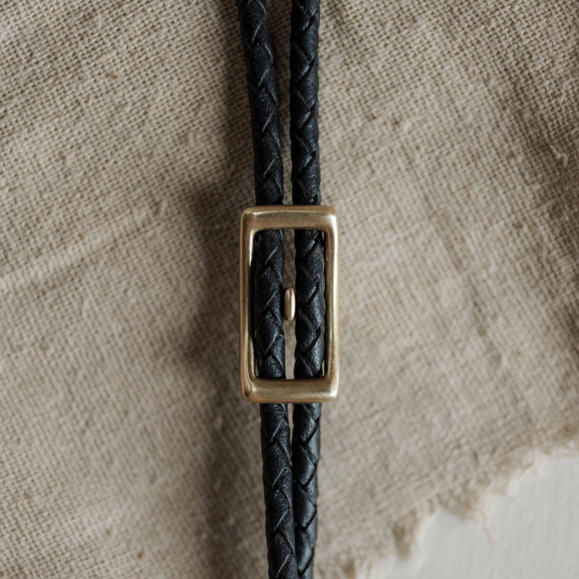 Black braided leather strap with a brass buckle on a textured beige background
