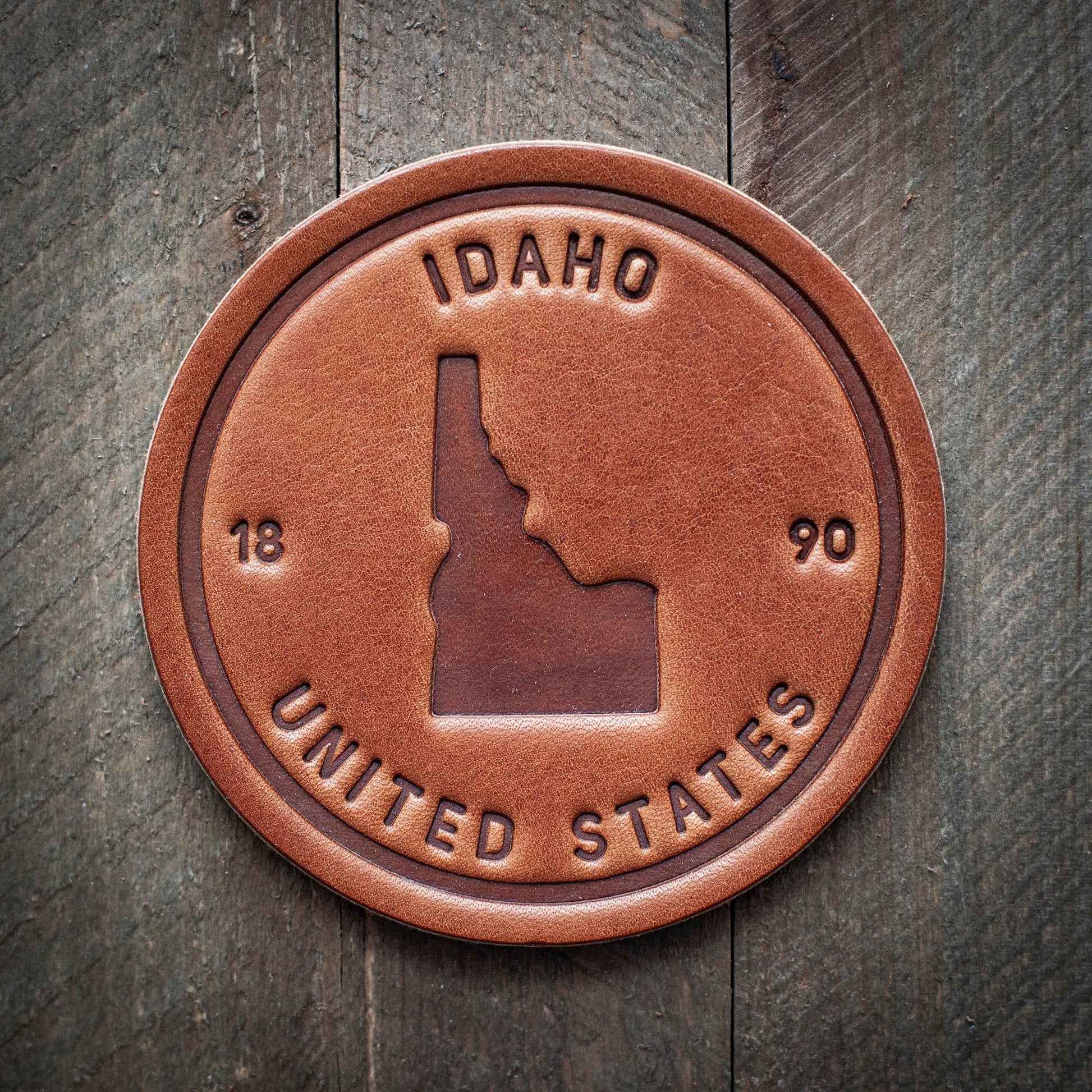 A round brown leather coaster featuring an embossed Idaho state silhouette, the words 'IDAHO' and 'UNITED STATES,' and the numbers '18' and '90,' shown on a dark rustic wood background.