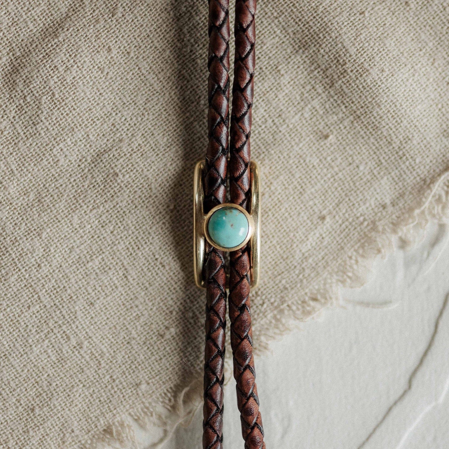 Brown braided leather bolo tie with a turquoise stone and gold clasp on a textured beige background.