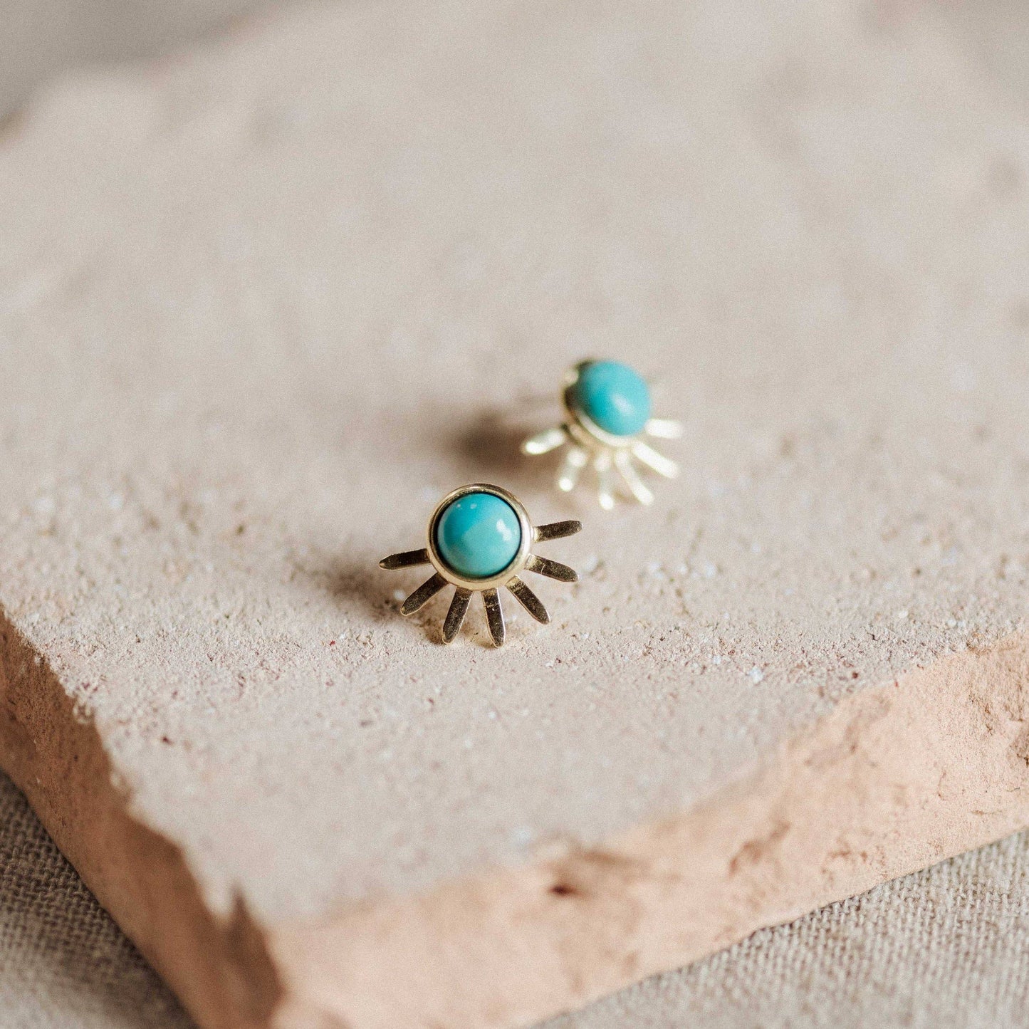 A pair of gold-toned stud earrings with round turquoise blue centers and sunburst rays, displayed on a textured beige stone slab.