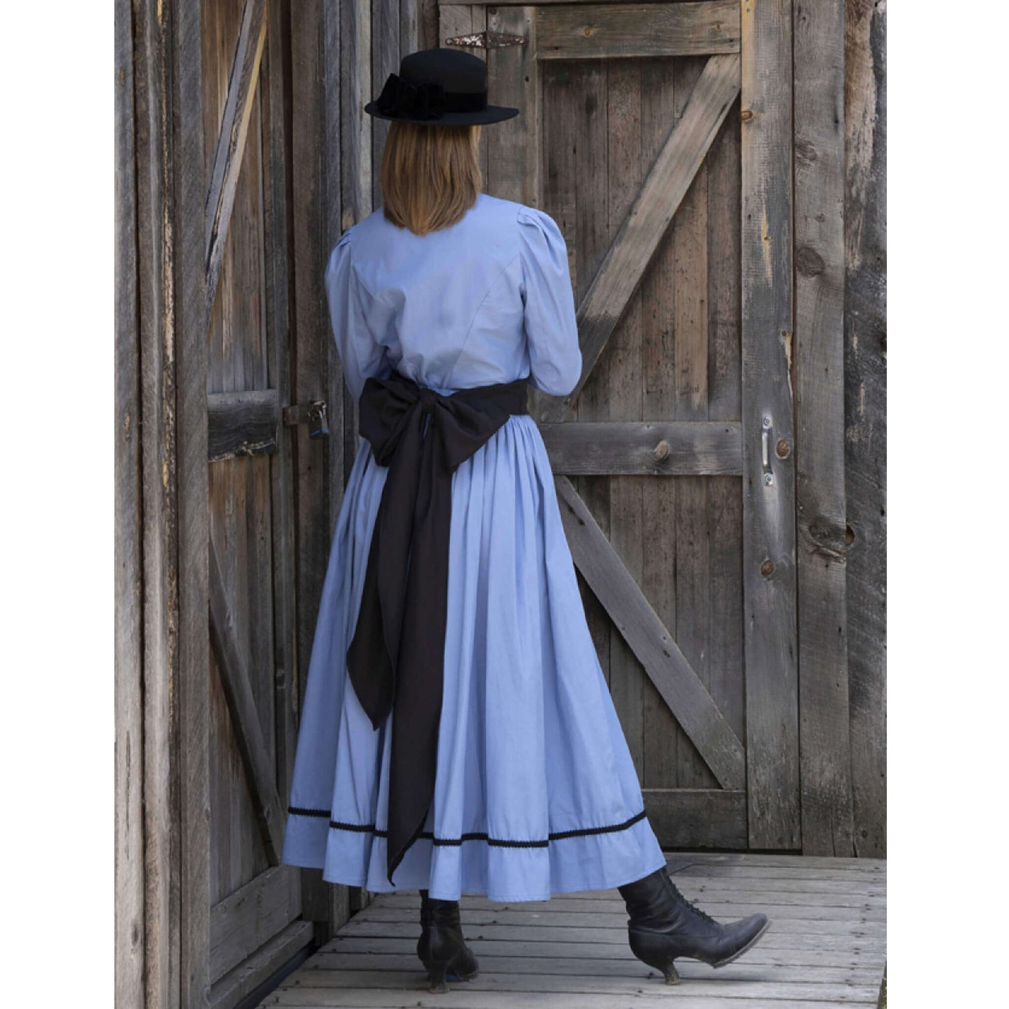 Back view of a woman in a Ladies Old West Bowman Outfit with a blue dress and black accents, standing near wooden doors.