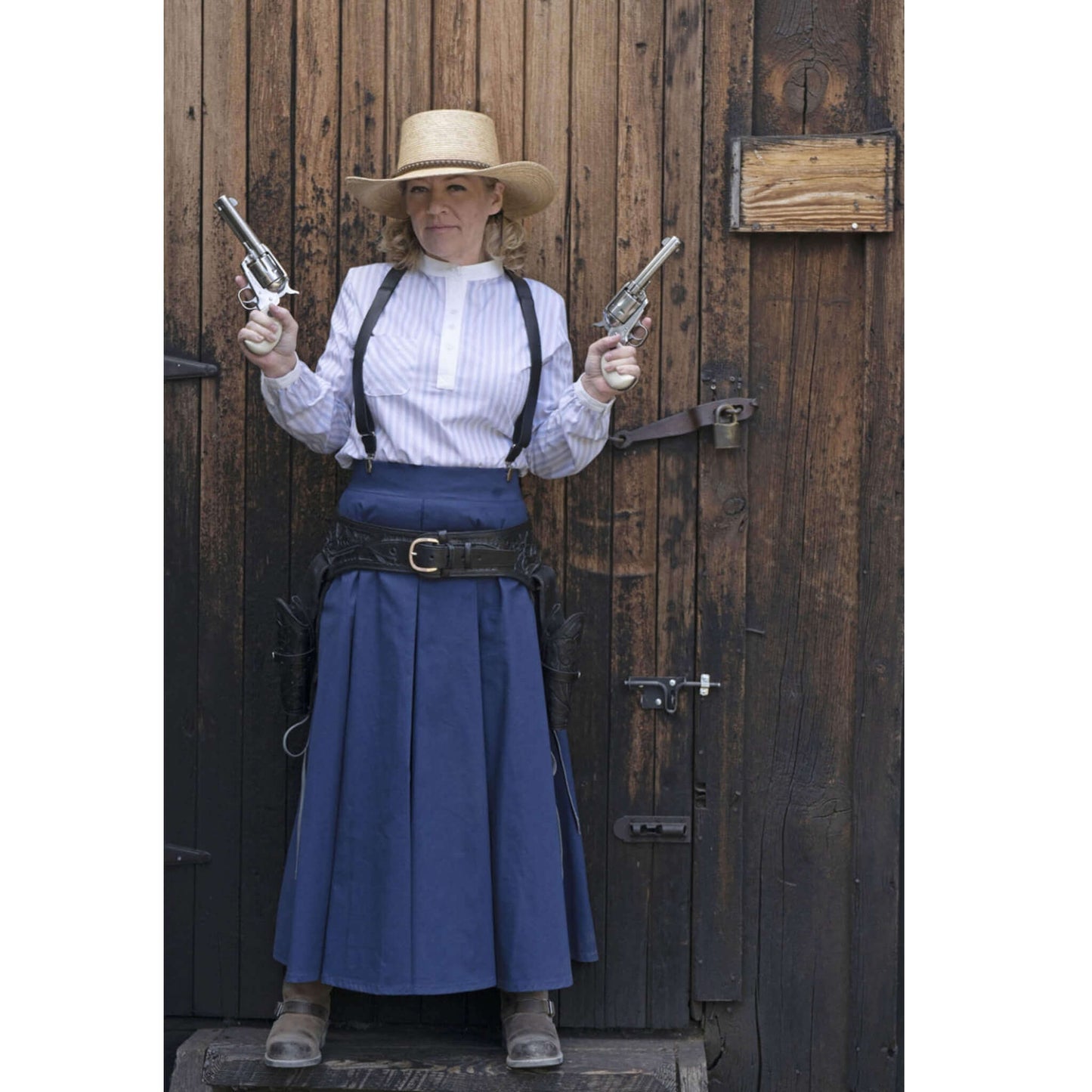 Woman in Old West attire holding revolvers, wearing a traditional blue skirt and shirt, posed against a wooden backdrop.