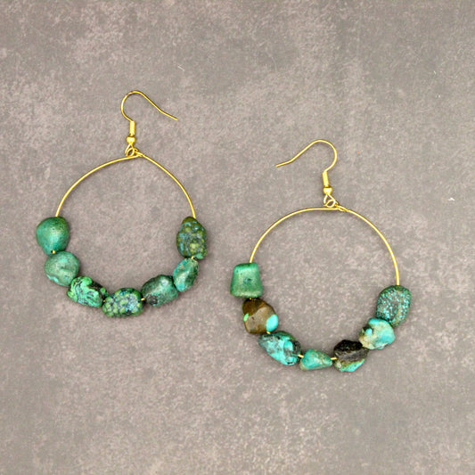 A pair of gold-tone hoop earrings with irregular green and turquoise stone beads, displayed on a textured gray background.