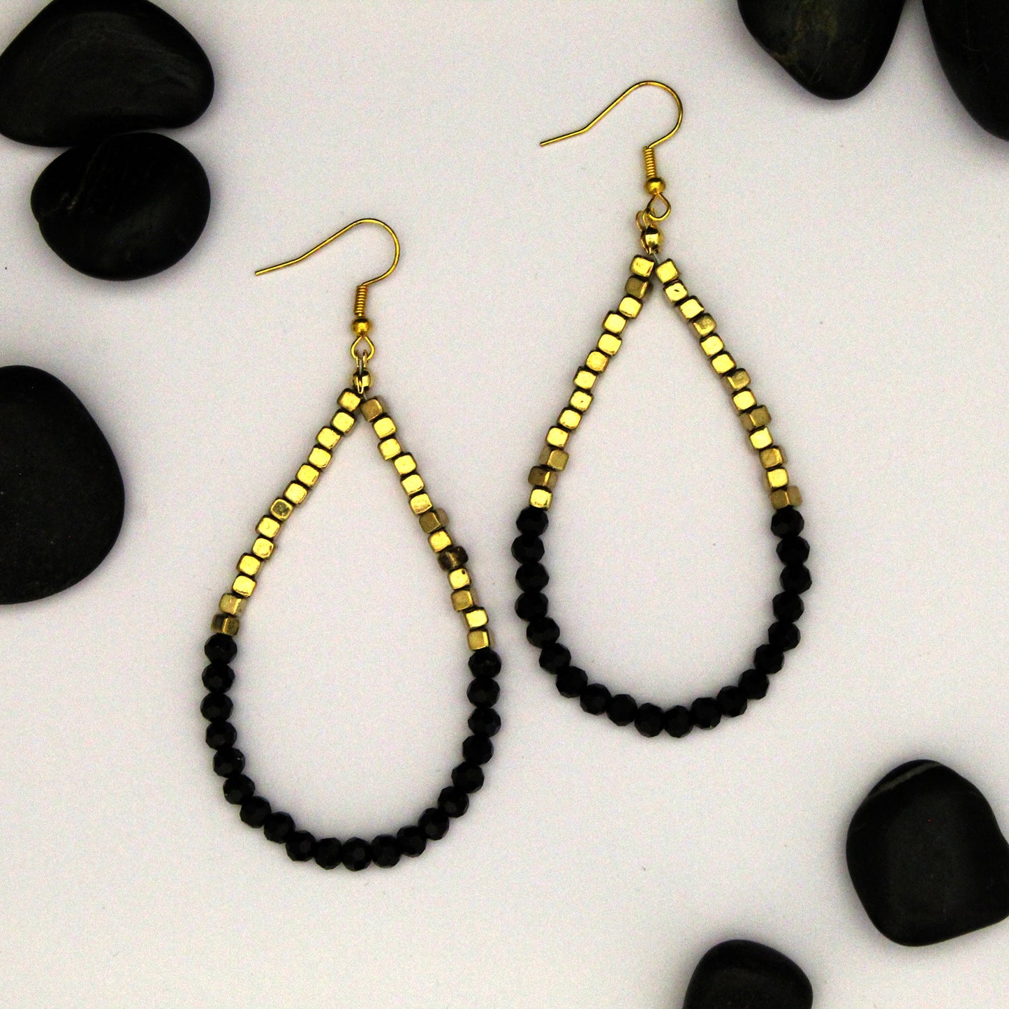 A pair of teardrop-shaped dangle earrings with faceted gold-tone top sections and black faceted bead loops, displayed on a white background with scattered black decorative stones.