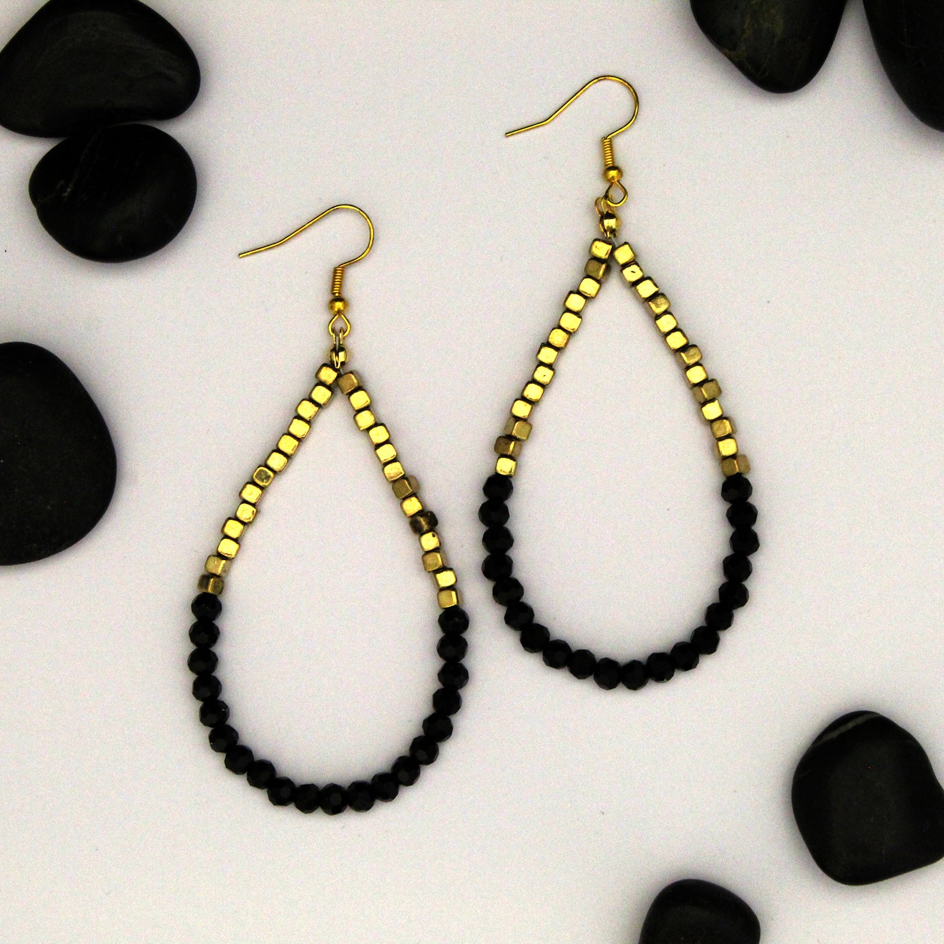 A pair of teardrop-shaped dangle earrings with faceted gold-tone top sections and black faceted bead loops, displayed on a white background with scattered black decorative stones.