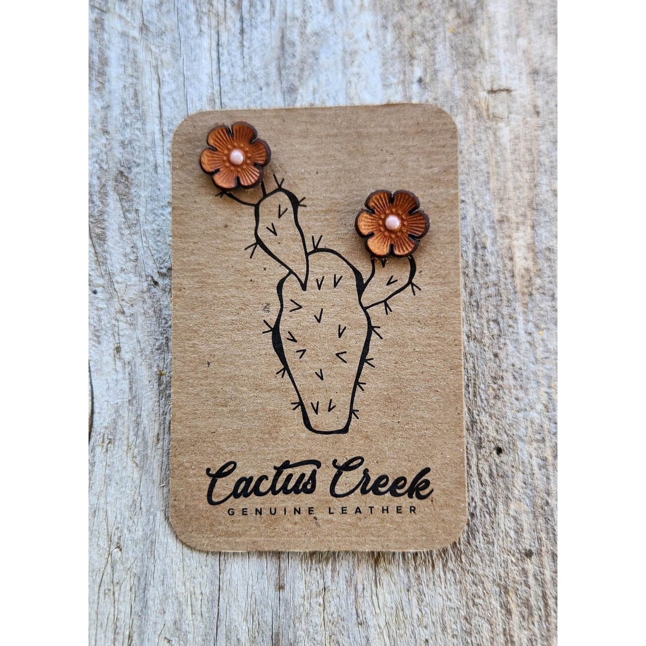 A pair of brown leather cactus-shaped stud earrings with small pearl-like centers, attached to a branded cardboard display card that reads 'Cactus Creek Genuine Leather,' photographed on a light weathered wood surface.