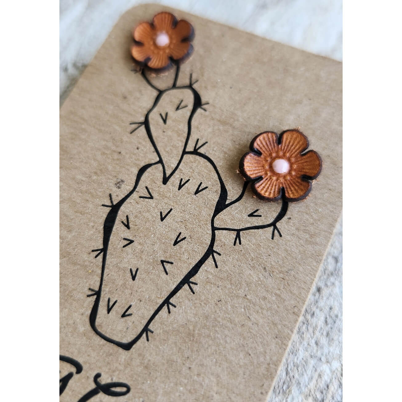 A pair of leather flower stud earrings with brown petals, dark brown thorns, and light pink centers, displayed on a brown kraft paper card with a cactus illustration.