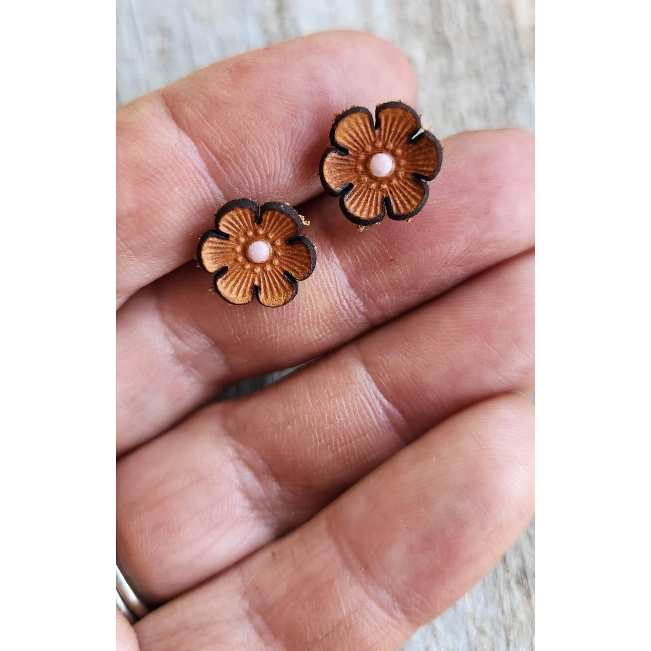A pair of small flower-shaped stud earrings with textured brown petals and pale pink centers, shown resting on a person's hand.
