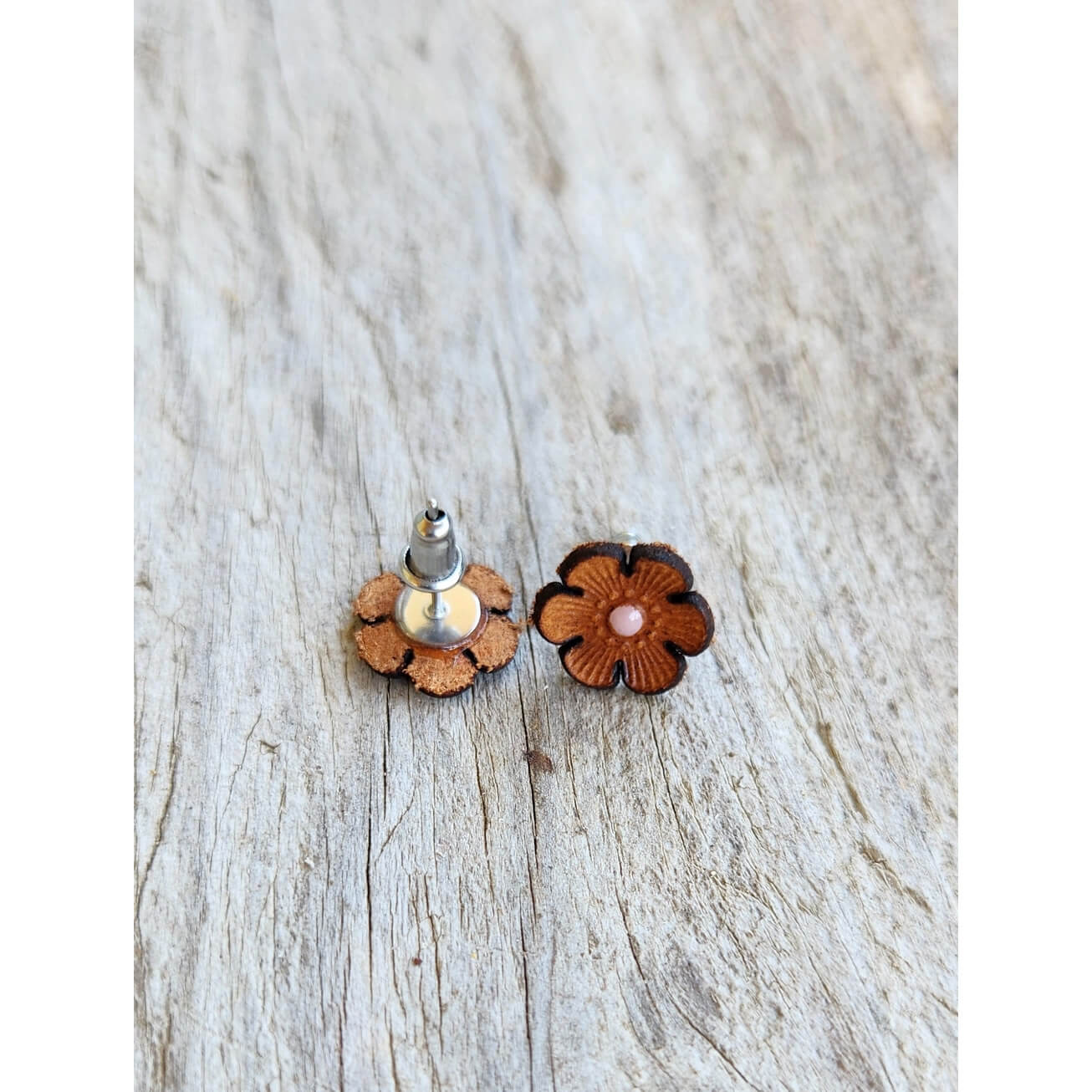 A pair of small flower-shaped stud earrings with layered brown wooden petals, one showing the silver-toned post and white pearl center, the other showing the front with a pink center, placed on a light weathered wood surface.