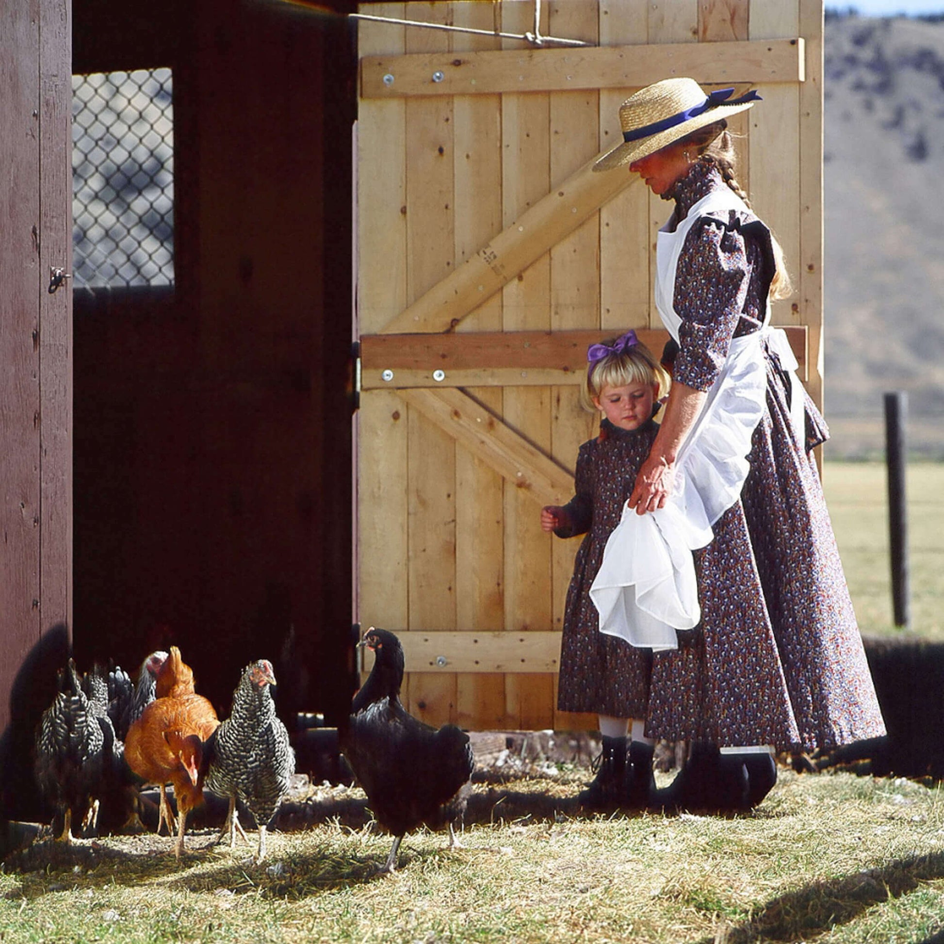 Woman and girl in traditional clothing with chickens near a barn, showcasing rural farm life and chicken feeding.