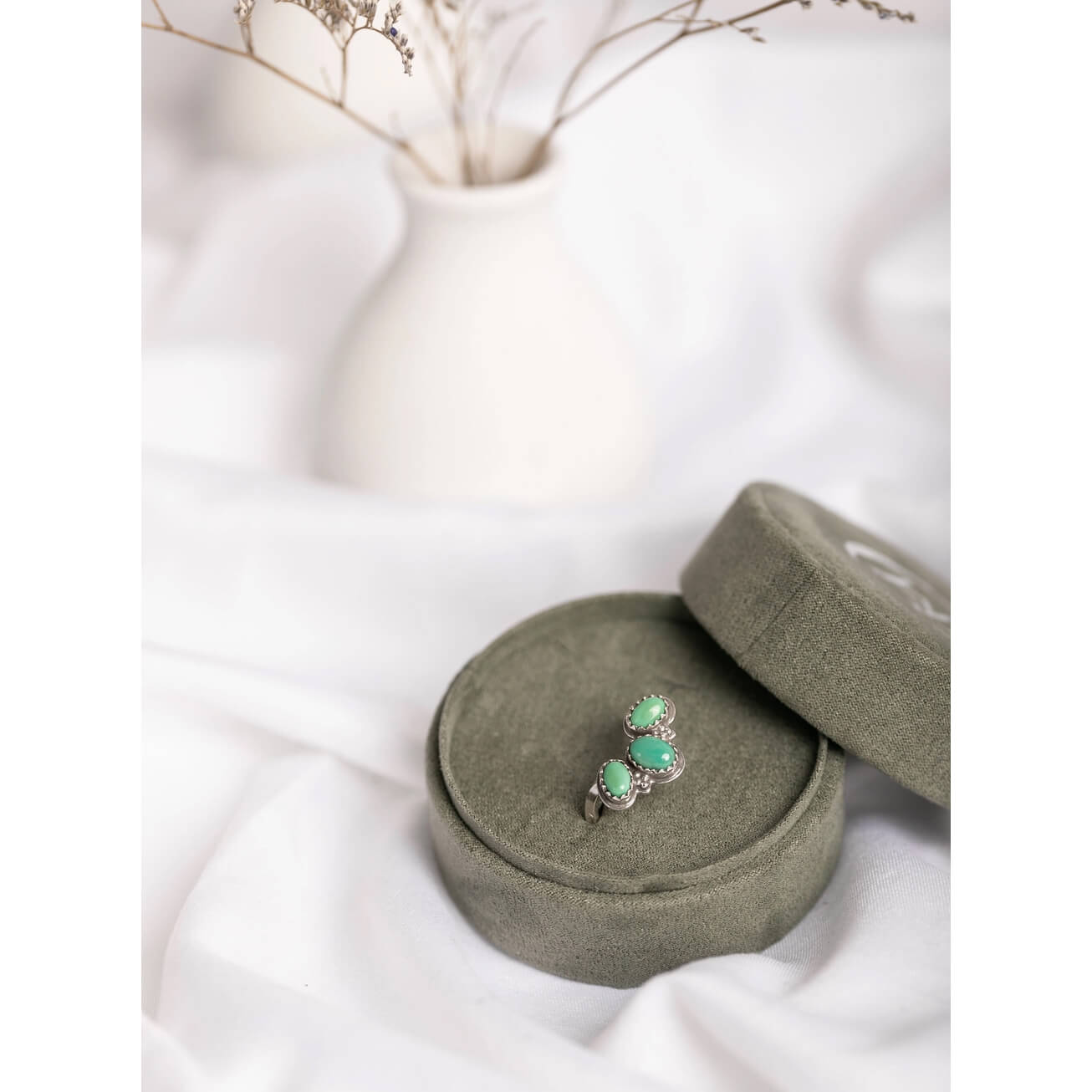 A sterling silver ring with three round turquoise stones, displayed on a round olive green velvet ring box on draped white fabric.