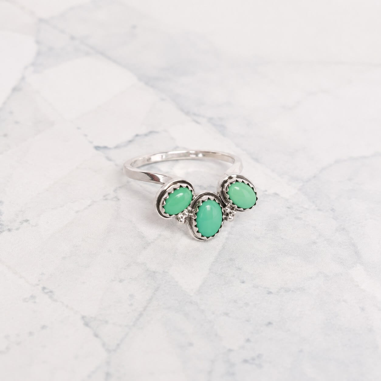 A silver ring featuring three oval-shaped turquoise stones, displayed on a light gray and white marble surface.