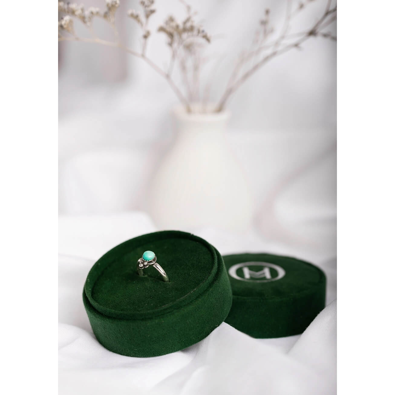 A silver ring with a round turquoise-colored stone sits in an open dark green velvet jewelry box, with a second closed box beside it on white fabric. A soft-focus white vase with dried stems is blurred in the background.