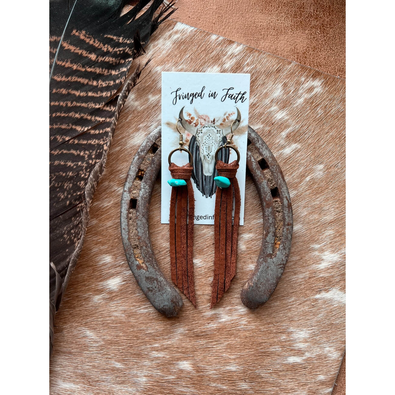 A pair of bohemian-style earrings displayed on a white backing card labeled 'Faith'. The earrings feature a silver-toned skull, brown leather fringe, and turquoise blue beads, arranged on a rustic brown and white cowhide surface with a feather and horseshoe as props.