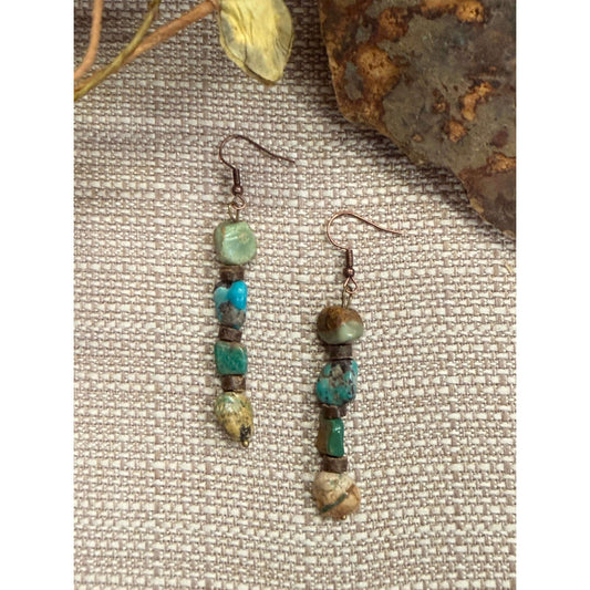 A pair of handmade dangle earrings with turquoise and green stone beads on copper-toned hooks, displayed on a textured beige fabric background with rustic decorative props.