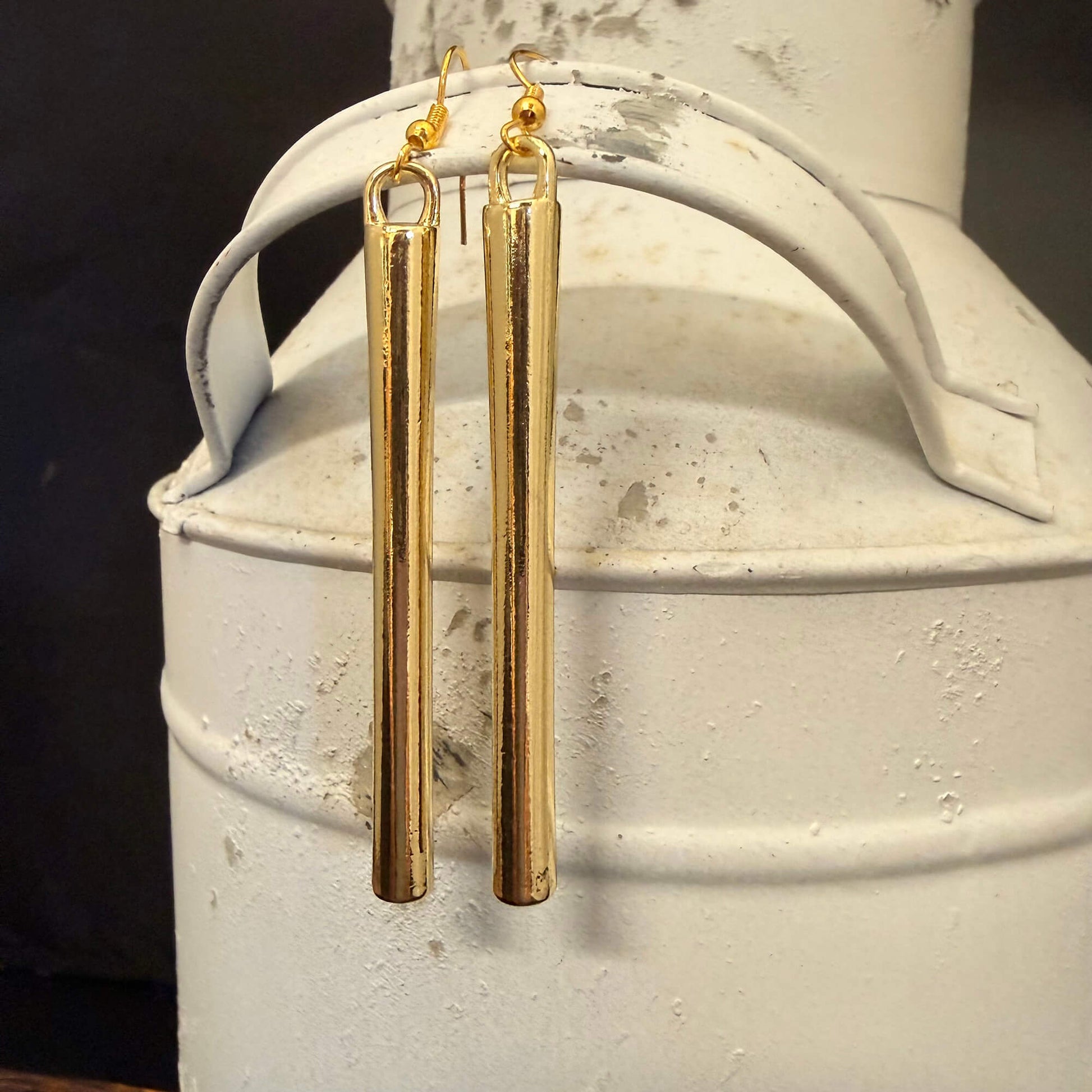 A pair of gold-toned drop earrings with elongated cylindrical bar charms, hanging from a rustic white metal canister against a dark background.