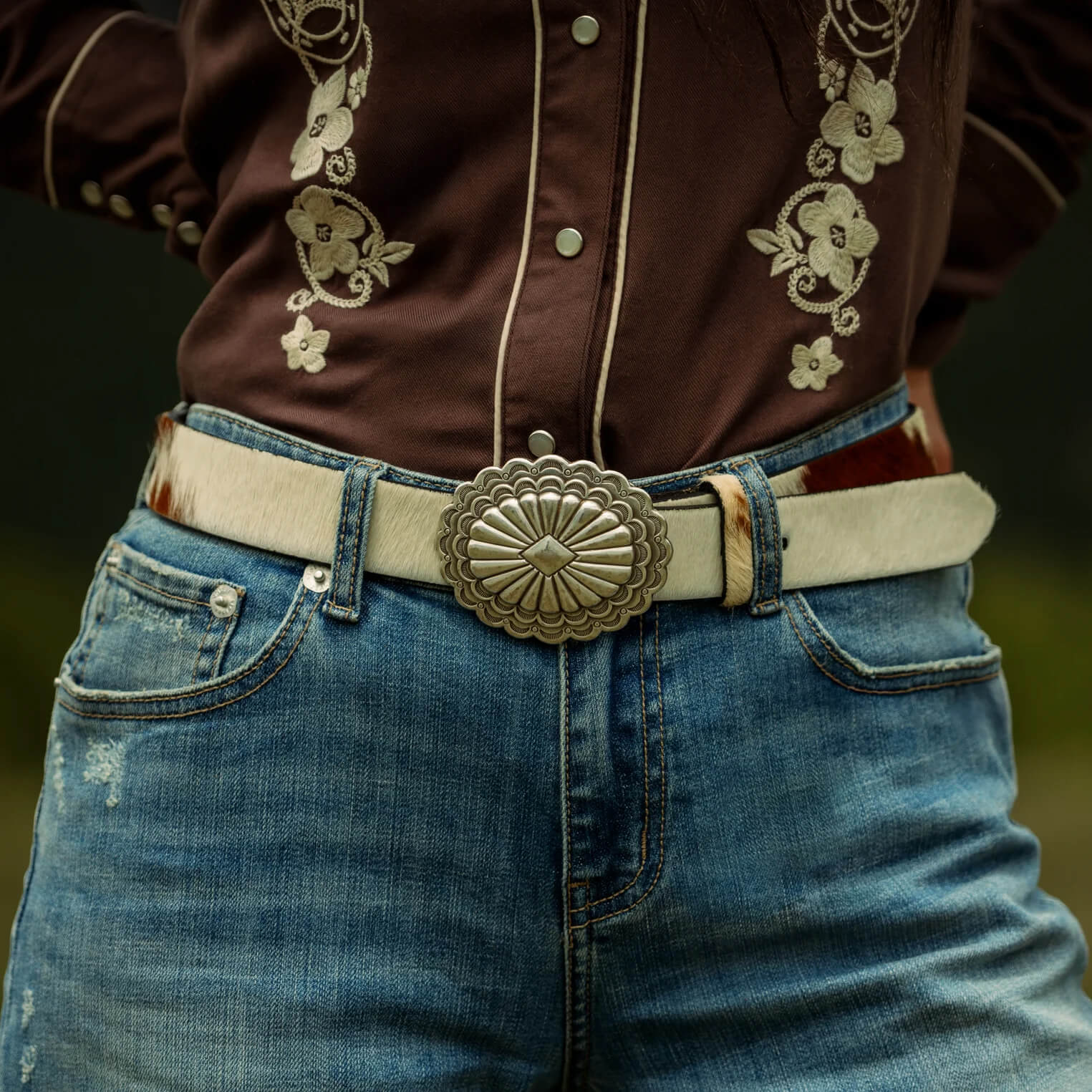 A close-up of a person wearing a brown embroidered western-style shirt, blue jeans, and a cream-colored belt with a large ornate silver-toned buckle.