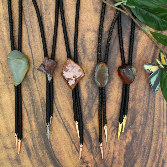 A selection of unique, handcrafted stone bolo ties laid out on a wooden surface, each with a different gemstone on a thick black leather cord with metal tips.