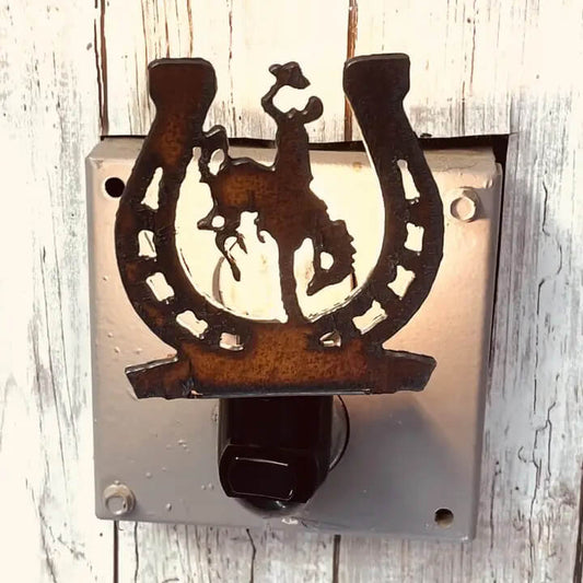 A rustic metal horseshoe-shaped night light featuring a cowboy silhouette, mounted on a square metal backplate with a black outlet insert, set against a whitewashed wood background.