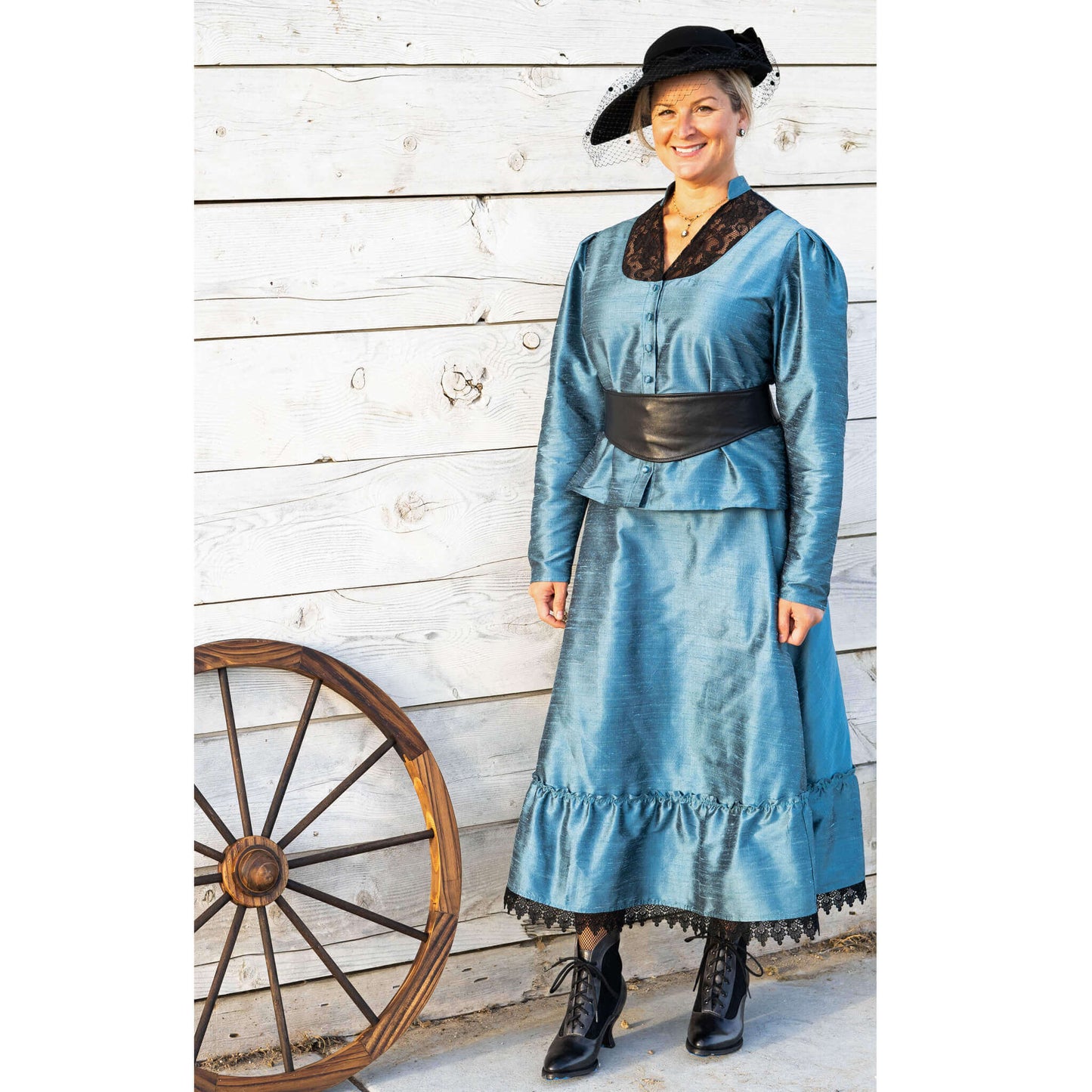 Woman in teal 1800's Old West outfit with black lace accents and hat, next to a wooden wagon wheel.