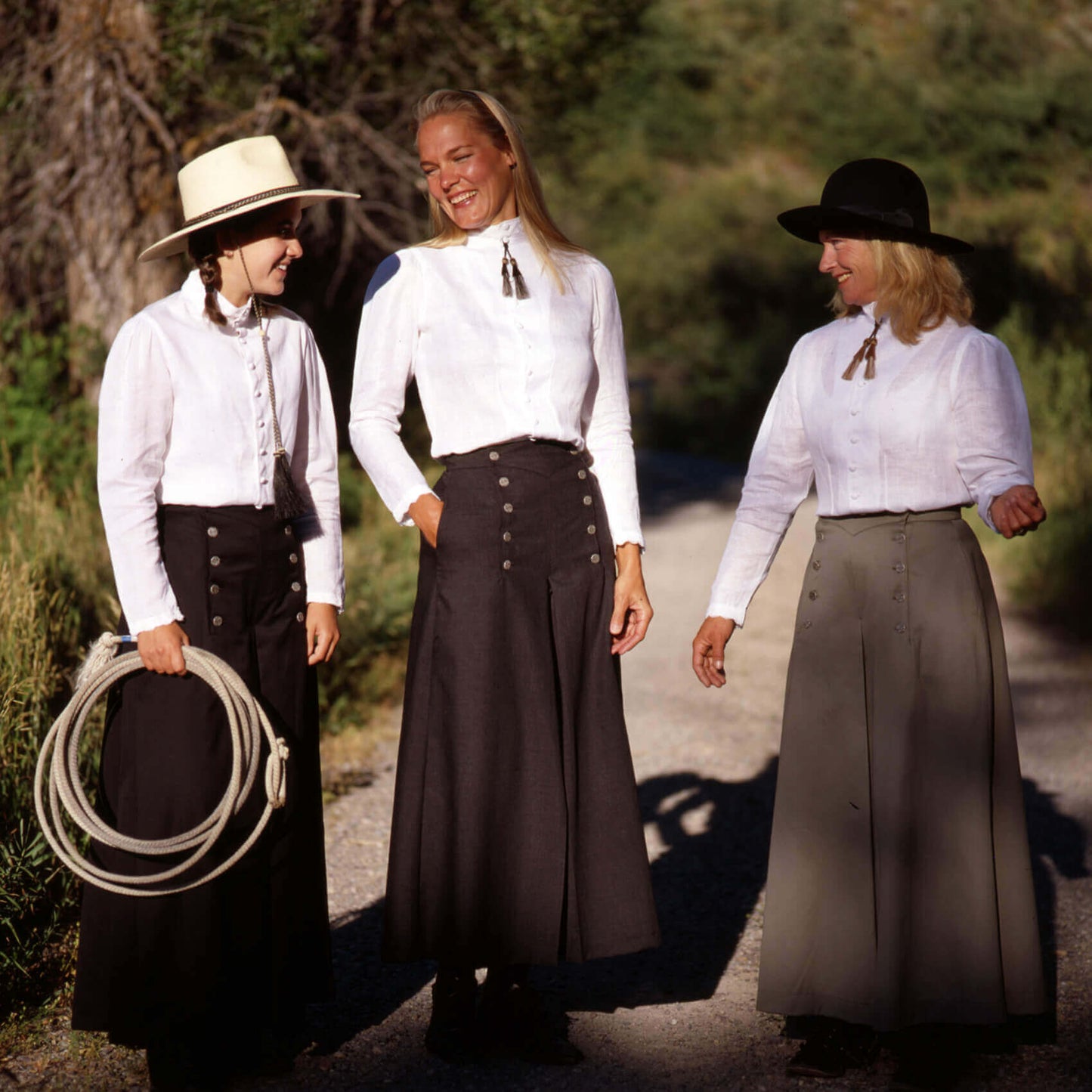 Three women in elegant 1800s-style blouses and long skirts, showcasing handmade lace details in a natural outdoor setting.