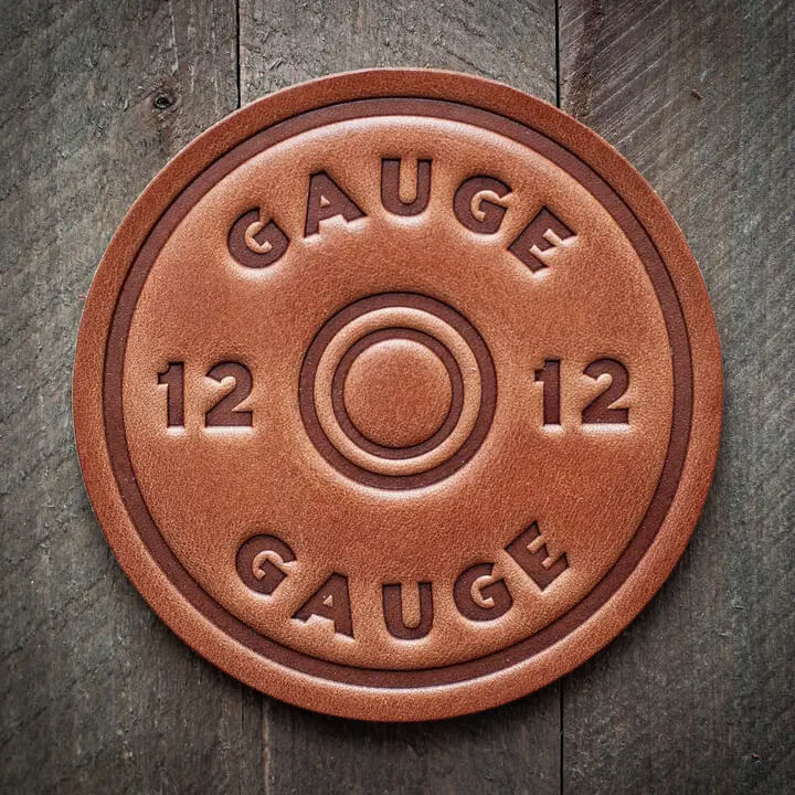 A round leather coaster embossed with the words 'GAUGE 12' and a central ring motif, photographed on a dark rustic wooden surface.