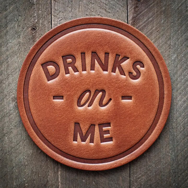 A round brown leather coaster with embossed text reading 'DRINKS on ME' in a dark brown tone, placed on a rustic dark wood surface.