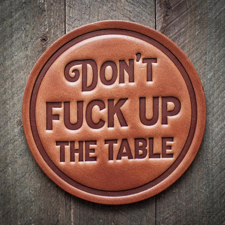 A round brown leather coaster with embossed text reading 'DON'T FUCK UP THE TABLE' in a dark brown tone, displayed on a rustic dark wood background.