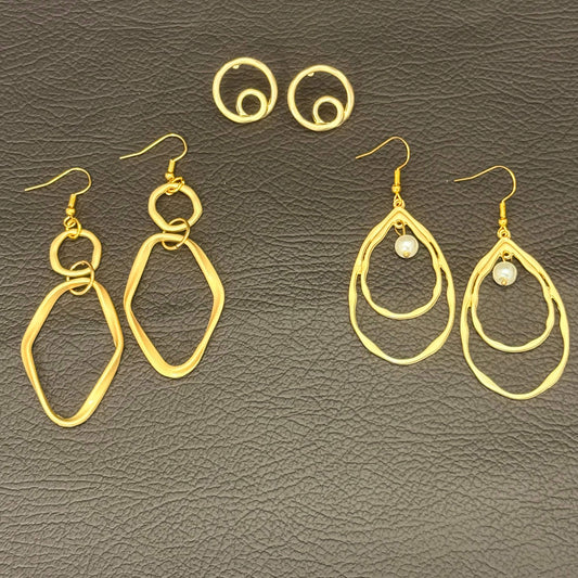Three pairs of gold-toned dangle earrings displayed on a dark textured leather background. The set includes two pairs of large abstract open-loop drop earrings and one pair of smaller hoop earrings, with one pair featuring a small white pearl accent.
