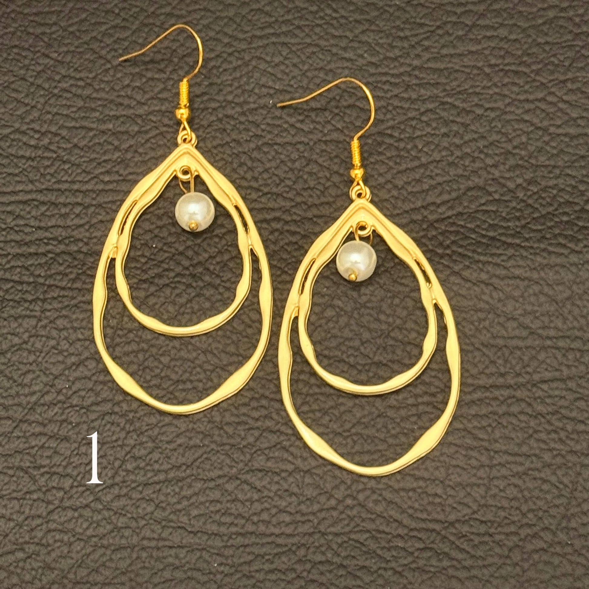 A pair of gold-toned dangle earrings with irregular double teardrop-shaped rings and a small white pearl at the center, displayed on a dark textured leather background.