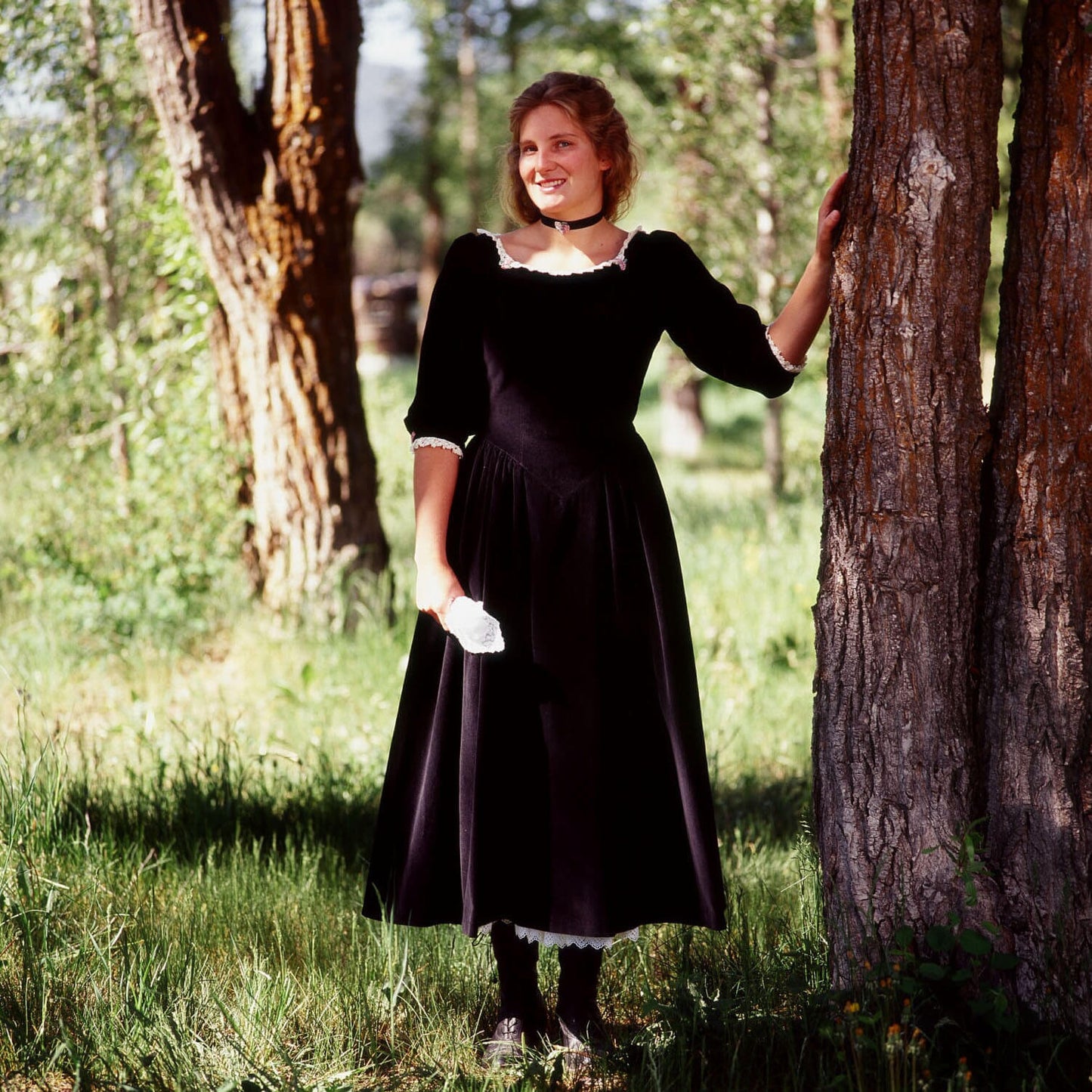 Woman in black velvet Maverick Queen vintage dress standing by a tree in a green forest setting.