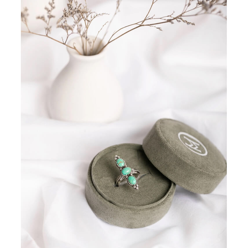 A silver ring with three round turquoise stones sits in an olive green velvet jewelry box, with the matching lid placed beside it on white fabric. A small white ceramic vase with dried stems is blurred in the background.