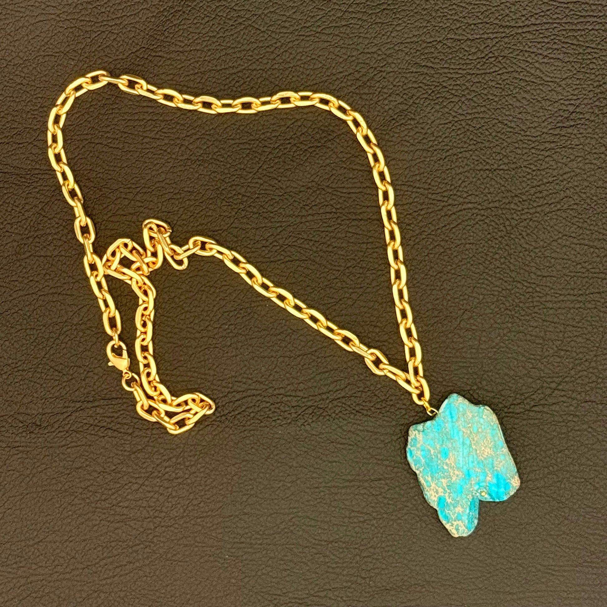 A gold chain necklace with a raw, irregular turquoise-blue stone pendant, displayed on a dark brown textured leather background.