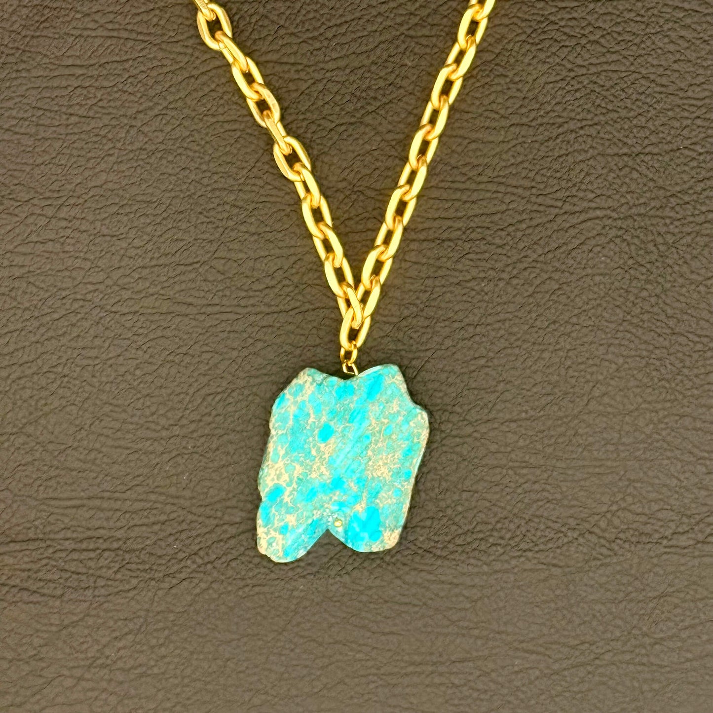 A gold chain necklace with a raw, irregular-shaped turquoise-blue stone pendant, displayed on a textured dark brown leather background.
