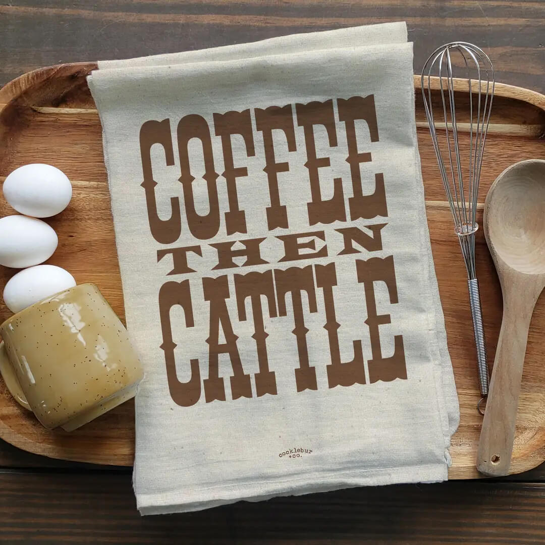 A beige linen tea towel with the words 'COFFEE THEN CATTLE' printed in large brown letters, laid flat on a wooden serving tray with eggs, a mug, a whisk, and a wooden spoon around it.