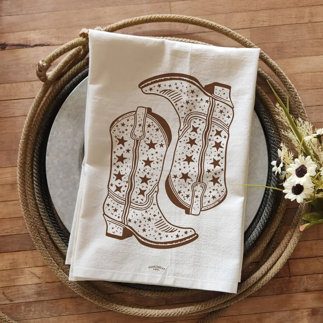 An off-white cotton tea towel featuring a brown printed graphic of cowboy boots with stars and dots, styled on a round wooden tray with a rope border and small white flowers on a wooden floor.