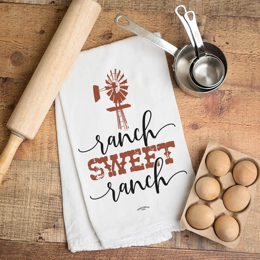 A white kitchen towel with a rustic 'ranch sweet ranch' design in black and terracotta red, laid flat on a wooden table alongside a wooden rolling pin, metal measuring cups, and a carton of eggs.
