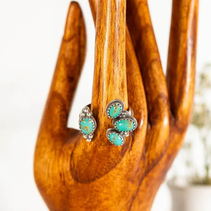 A Silver Ring with oval turquoise stones, displayed on a polished wooden hand stand against a soft white background.