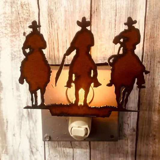 A rustic metal night light featuring three cowboy silhouettes on horseback, mounted on a wall plug against a weathered wood background. The center figure is illuminated by a warm light, casting a golden glow.