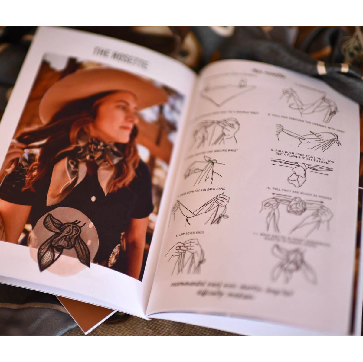 An open book showing a fashion illustration page with a woman wearing a hat and scarf, alongside black line drawings of hand poses. The pages are photographed on a soft, dark fabric background.