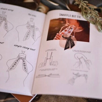 An open book or booklet showing illustrated tie-tying instructions, including line drawings of knot methods and a small inset photo labeled 'SNAFFLE BIT TIE.' The pages are photographed on a styled surface with decorative leaves and fabric in the background.