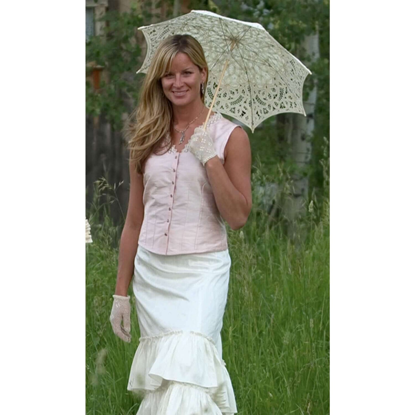 Elegant woman in a pink silk corset cover camisole with lace detail, holding a decorative parasol in a grassy setting.