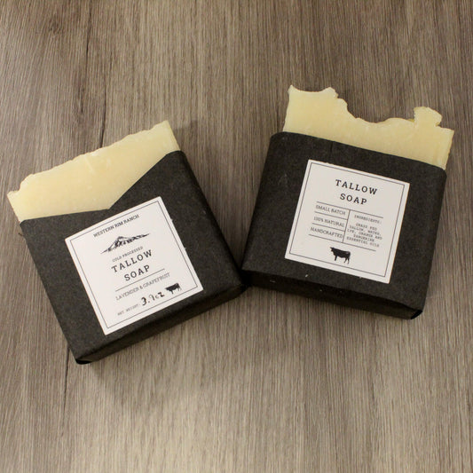 Two bars of Tallow Soap by Western Farm & Ranch, wrapped in dark charcoal paper with white labels, resting on a gray wood-textured surface.