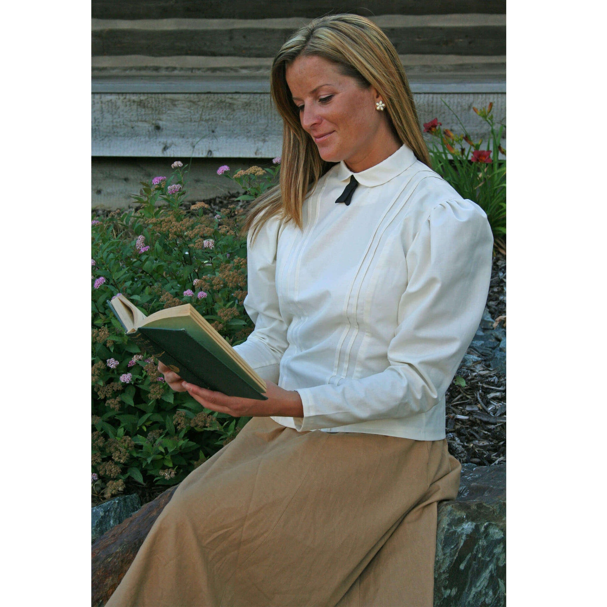 Woman in a Schoolmarm Country Blouse, reading a book outdoors, surrounded by flowers.