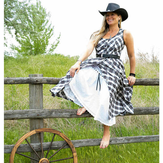 Woman in a plaid sleeveless western dress with eyelet petticoat, sitting on a fence in a summer setting.